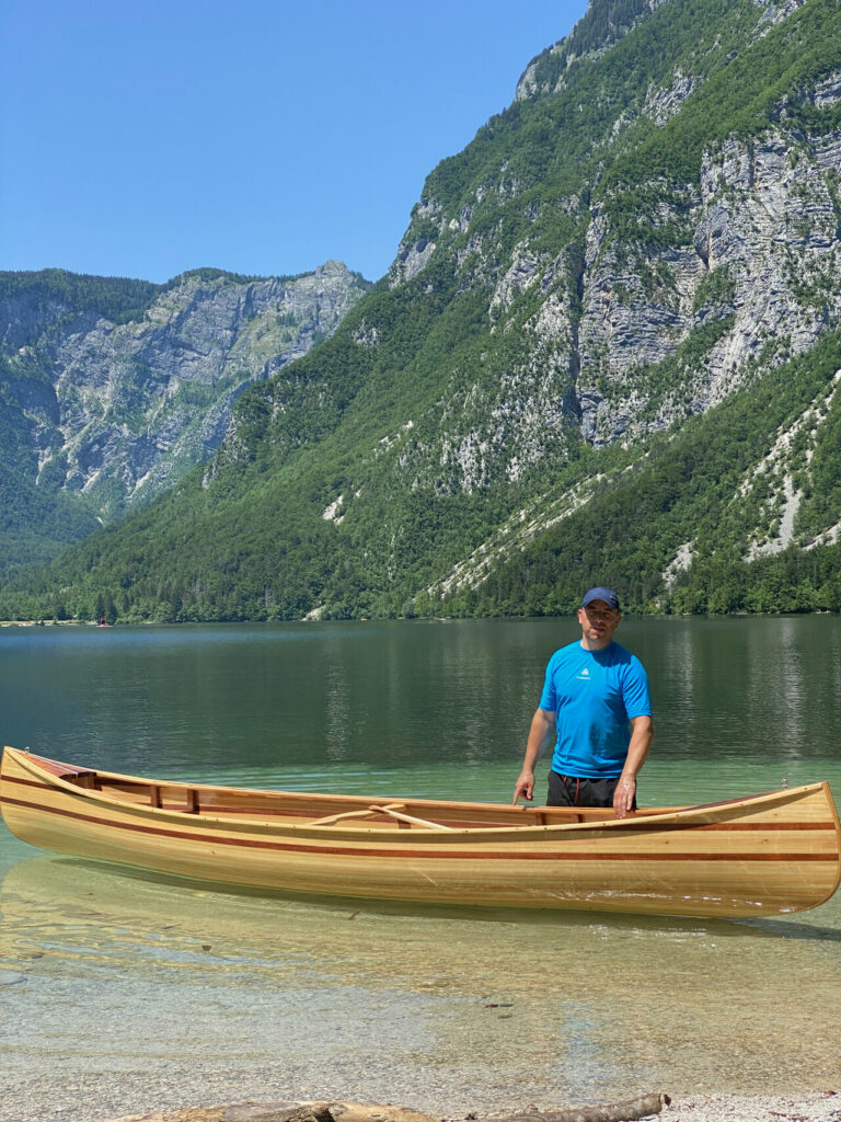 Jernej mit seinem selbst gebauten Holzkanu am See vor bewaldeten Bergen.